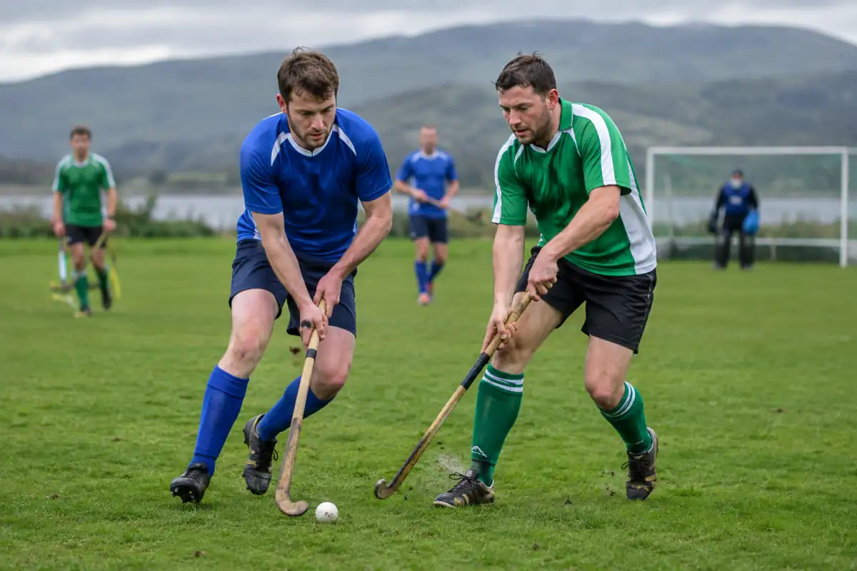 Deux joueurs disputent une balle de shinty au caman sur un terrain en herbe, avec un but visible en arrière-plan.