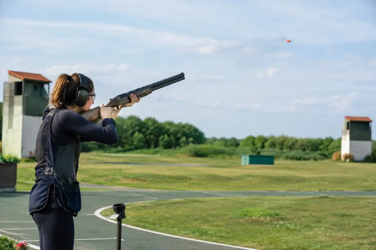 Un tireur équipé de lunettes et d’un casque suit un plateau d’argile sur un stand de skeet en plein air.