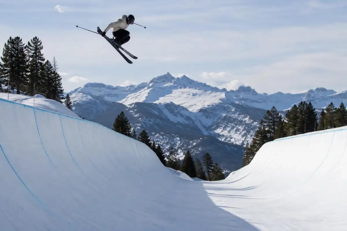 Un skieur de ski halfpipe s’élance au-dessus d’une paroi enneigée, skis twin-tip et casque, dans un snowpark alpin.