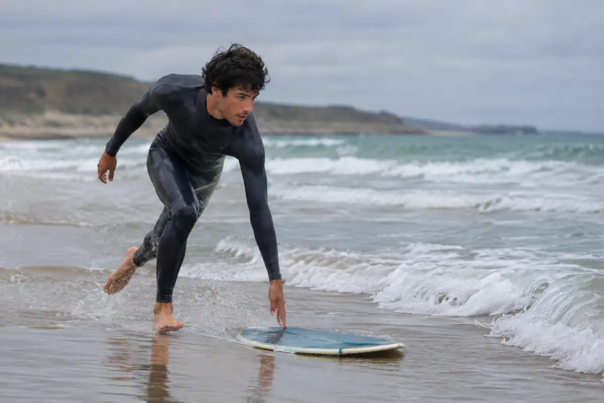 Skimboarder en combinaison qui lance sa planche sur le sable mouillé pour glisser vers une vague près du rivage.