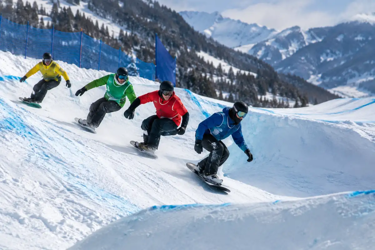 Des snowboardeurs s’élancent côte à côte sur un parcours de snowboard cross enneigé, entre virage relevé et bosses.