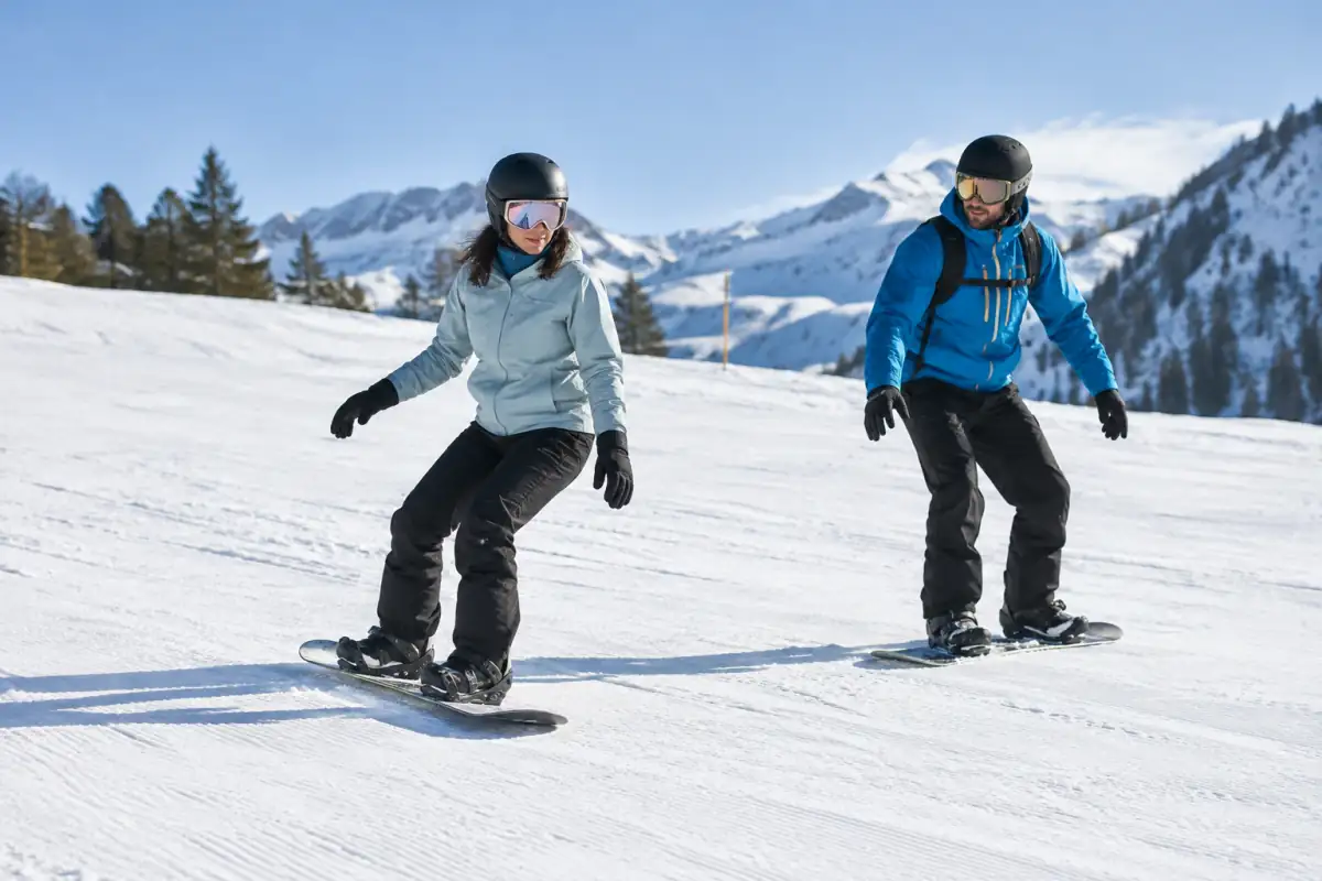 Débutant en snowboard travaillant l’arrêt sur la carre talons sur une piste verte, avec un moniteur à proximité.