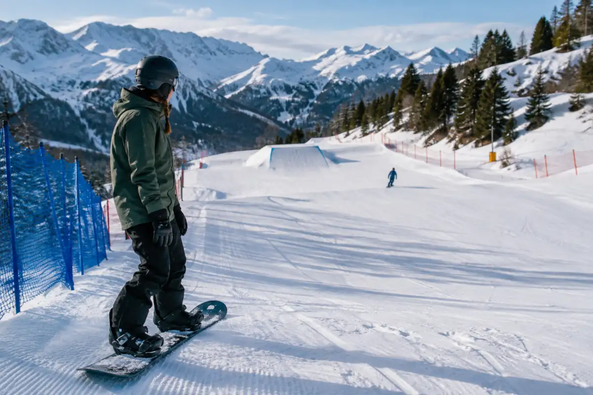 Un snowboardeur attend sur le bord d’un snowpark que la zone de réception du saut soit libre avant de s’élancer.