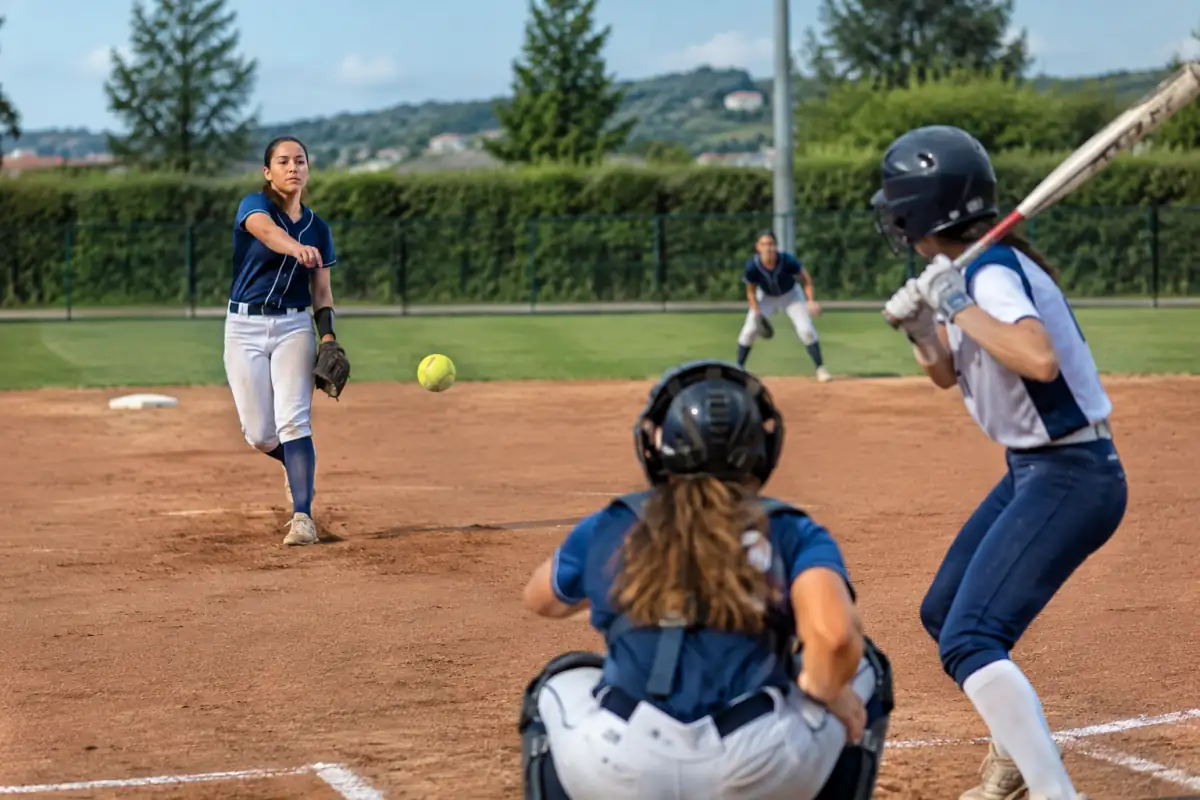 Une lanceuse de softball fastpitch effectue un lancer moulinet face à une frappeuse, avec receveuse protégée au marbre.