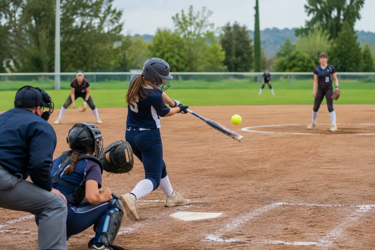 Une frappeuse en casque frappe une balle jaune lors d’un match de softball, avec le receveur protégé derrière le marbre.