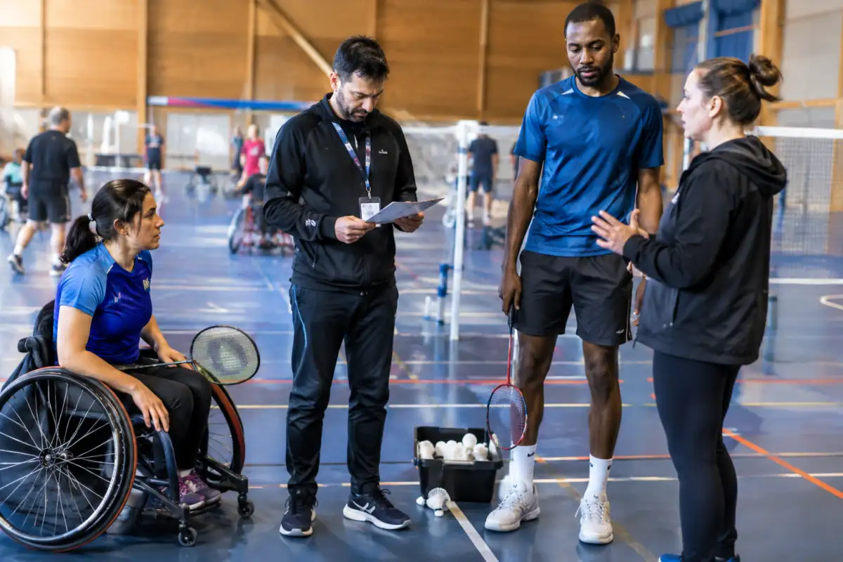 Un arbitre vérifie la feuille de score d’un match de badminton adapté tandis qu’un entraîneur échange avec un athlète.