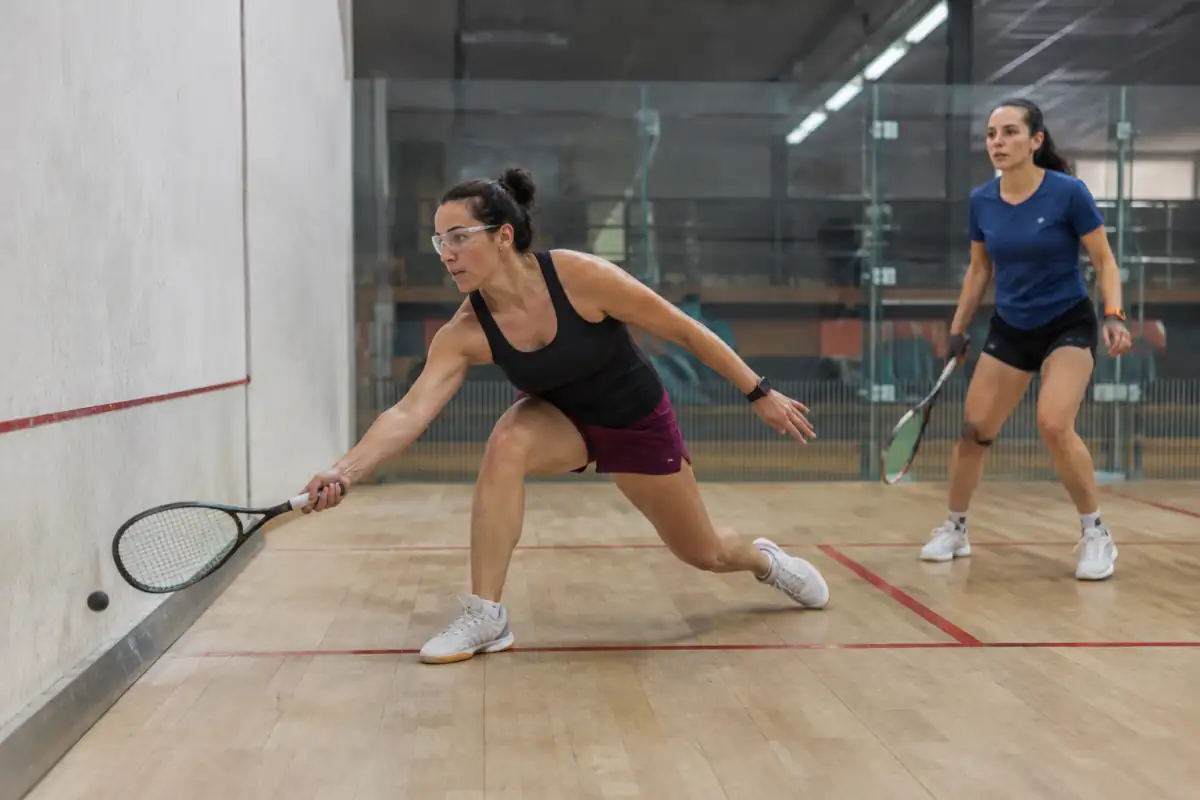 Deux joueurs de squash en salle échangent sur un court, l’un en fente basse avec raquette et lunettes de protection.