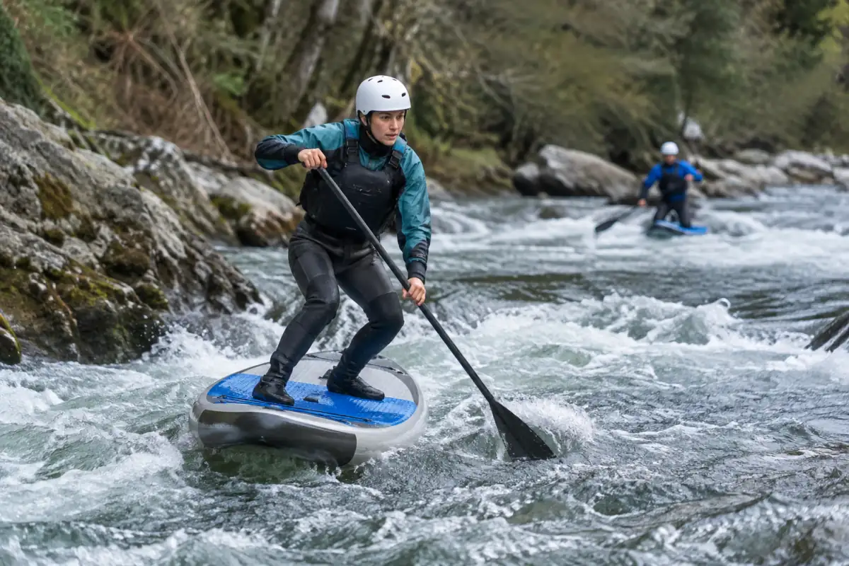 Pratiquant de stand up paddle en eaux vives debout sur une planche adaptée, pagaie en main, casque et aide à la flottabilité.