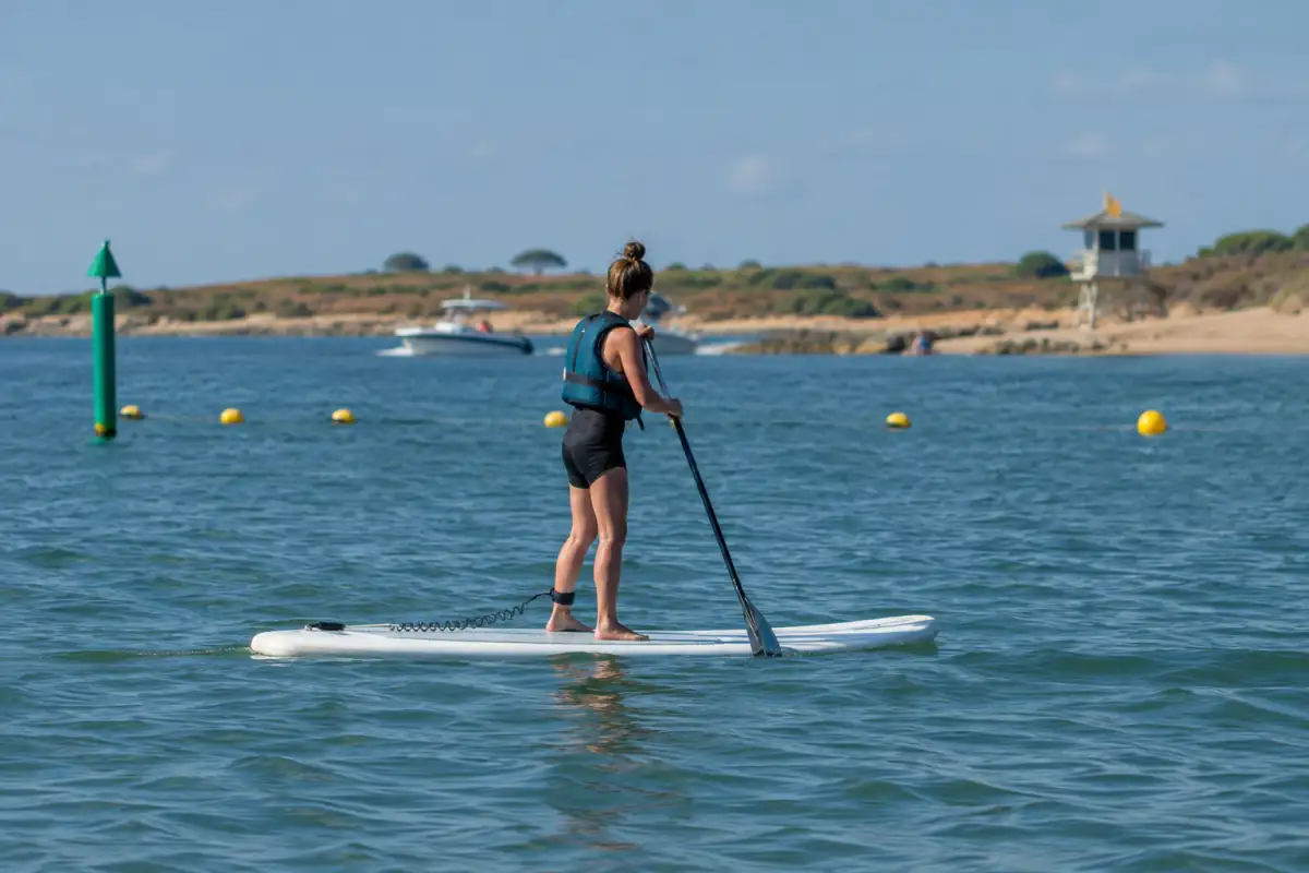 Un pratiquant en paddle, équipé d’un gilet, traverse un chenal côtier balisé en gardant ses distances avec les bateaux.