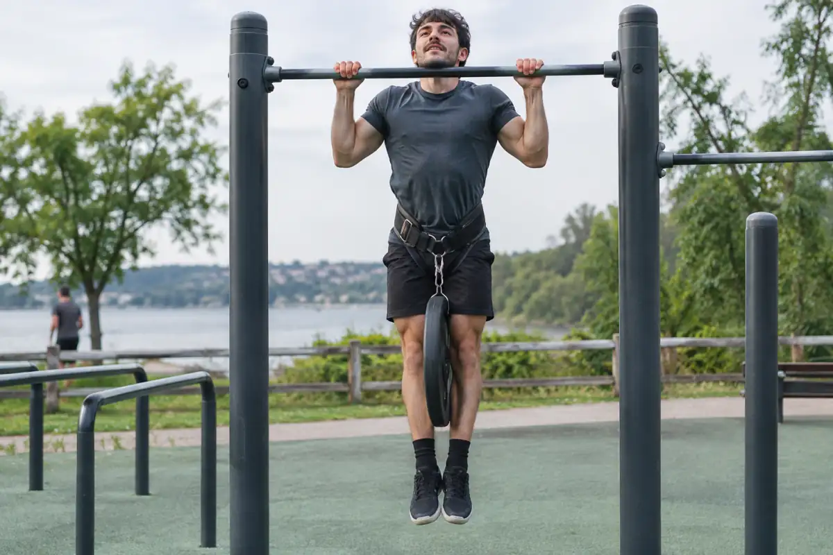 Athlète de streetlifting réalisant une traction stricte lestée sur une barre de parc, avec un disque suspendu à sa ceinture.