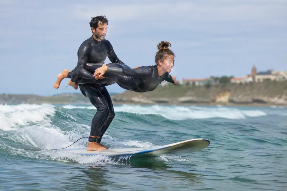 Deux surfeurs en combinaison réalisent un porté maîtrisé sur une longue planche de surf tandem, au-dessus d’une vague de l’Atlantique.