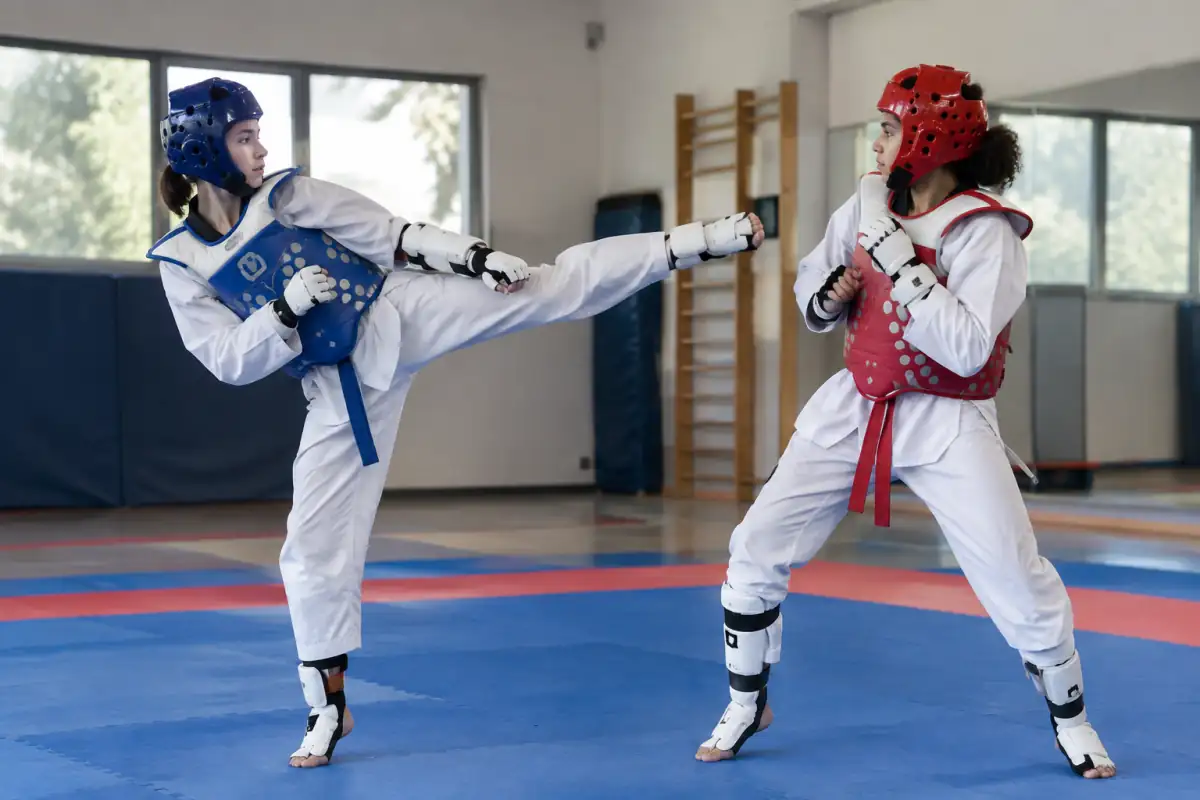 Deux taekwondoïstes en dobok s’entraînent au combat dans une salle, avec protections et coup de pied dirigé vers le plastron.