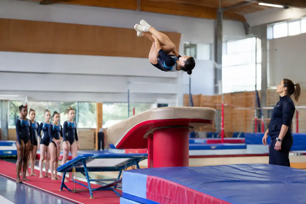 Gymnaste de TeamGym en plein saut sur mini-trampoline, sous le regard de son équipe et de l’entraîneur.