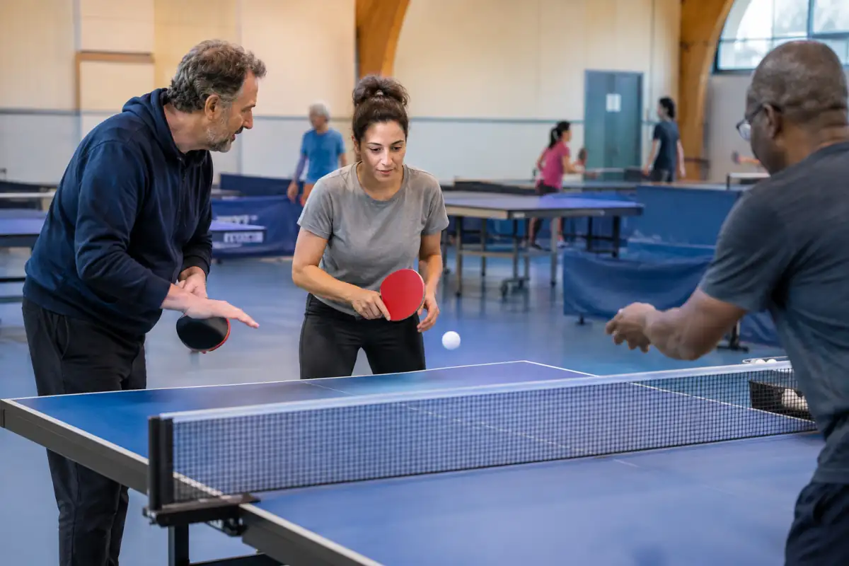 Un adulte débutant découvre le tennis de table en club, raquette en main, pendant un exercice guidé près d’une table réglementaire.