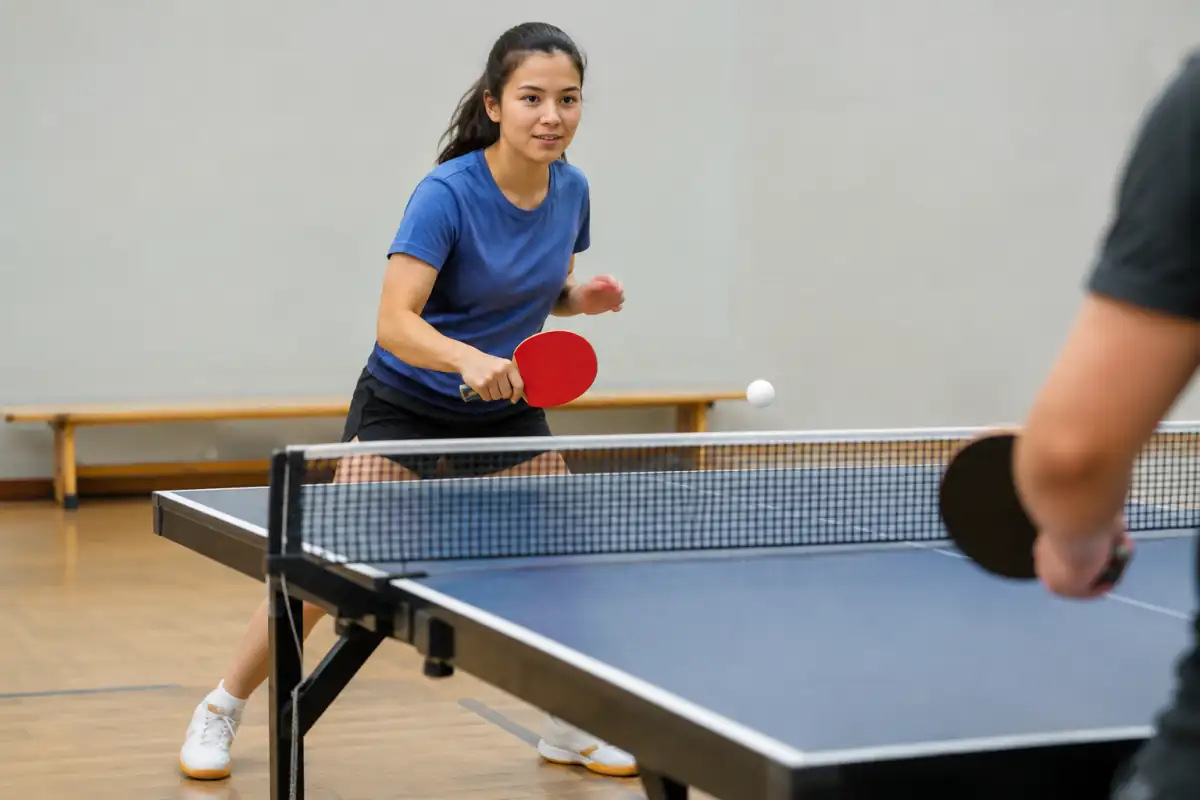 Débutant testant une raquette polyvalente de tennis de table près du filet, avec balle de 40 mm et chaussures de salle.