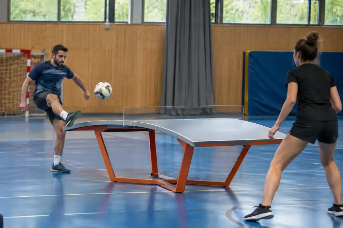 Deux joueurs échangent un ballon du pied au-dessus d’une table incurvée pendant une partie de teqball en salle.