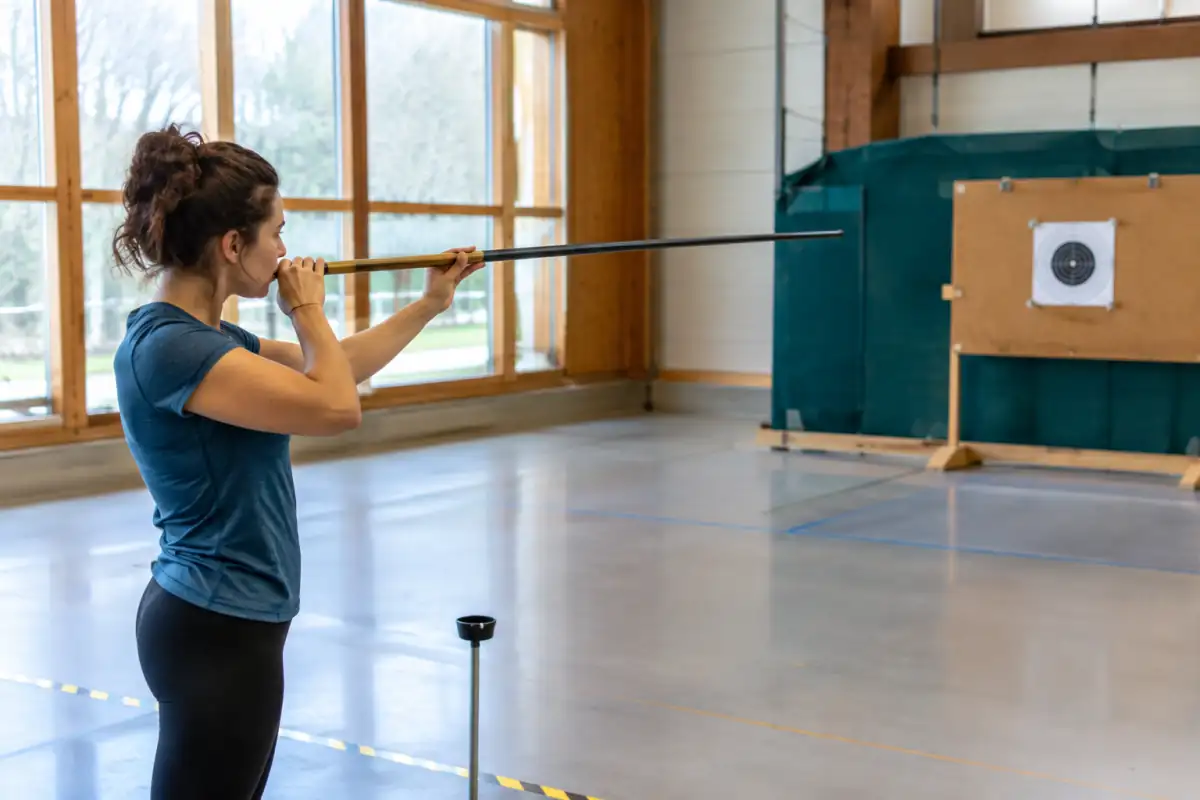 Une pratiquante de tir à la sarbacane souffle vers une cible installée au fond d’un pas de tir sécurisé en salle.
