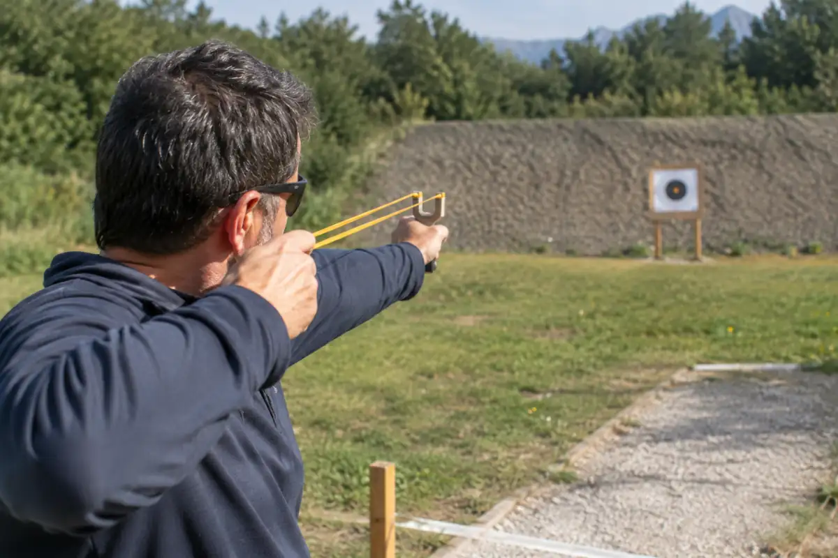 Un adulte portant des lunettes de protection vise une cible avec un lance-pierre sur un pas de tir sécurisé.