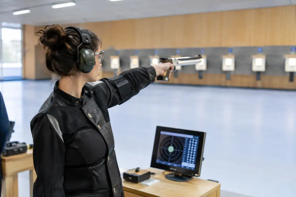 Une tireuse pratique le tir au pistolet à air comprimé à 10 mètres, debout face à une cible électronique dans un stand couvert.