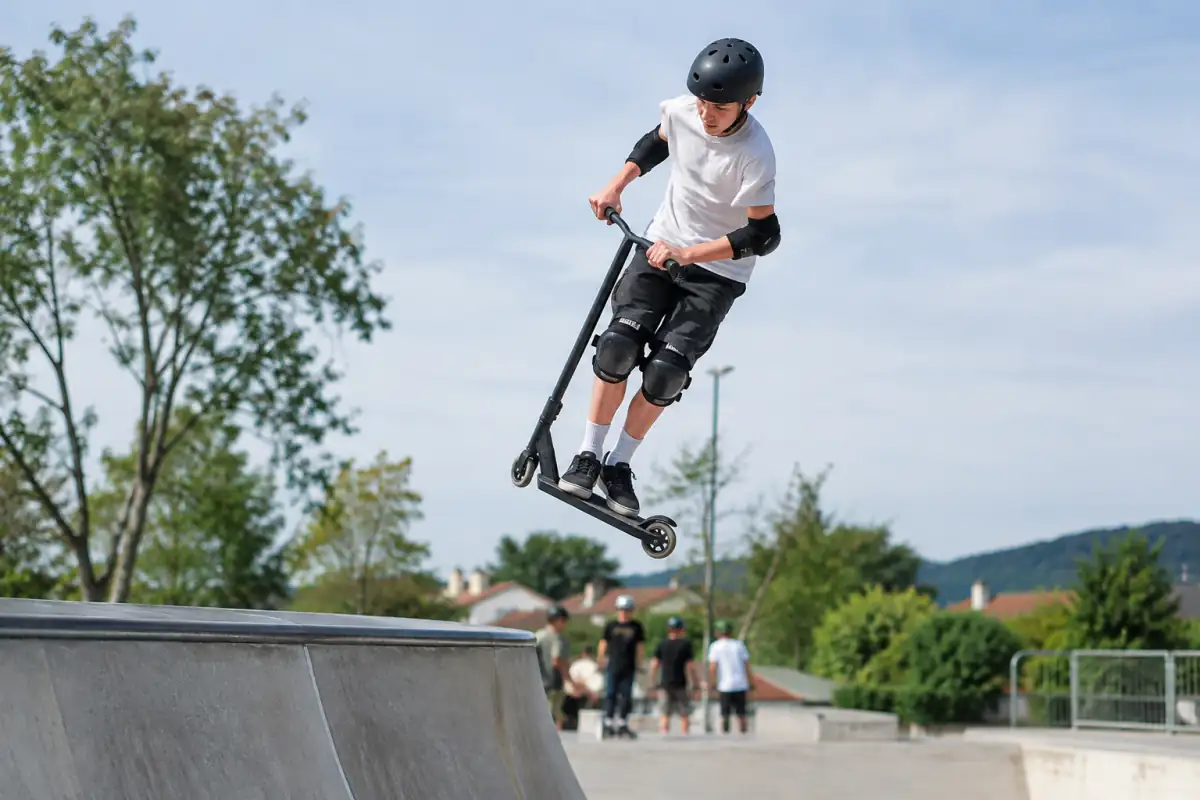 Un rider en trottinette freestyle park s’élance depuis un quarter-pipe dans un skatepark extérieur, casque et protections visibles.