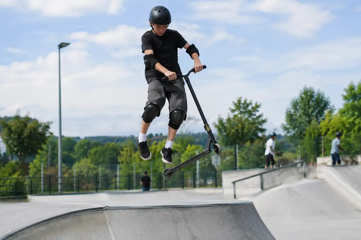 Un rider en trottinette freestyle réalise un tailwhip au-dessus d’une rampe dans un skatepark, avec casque et protections.