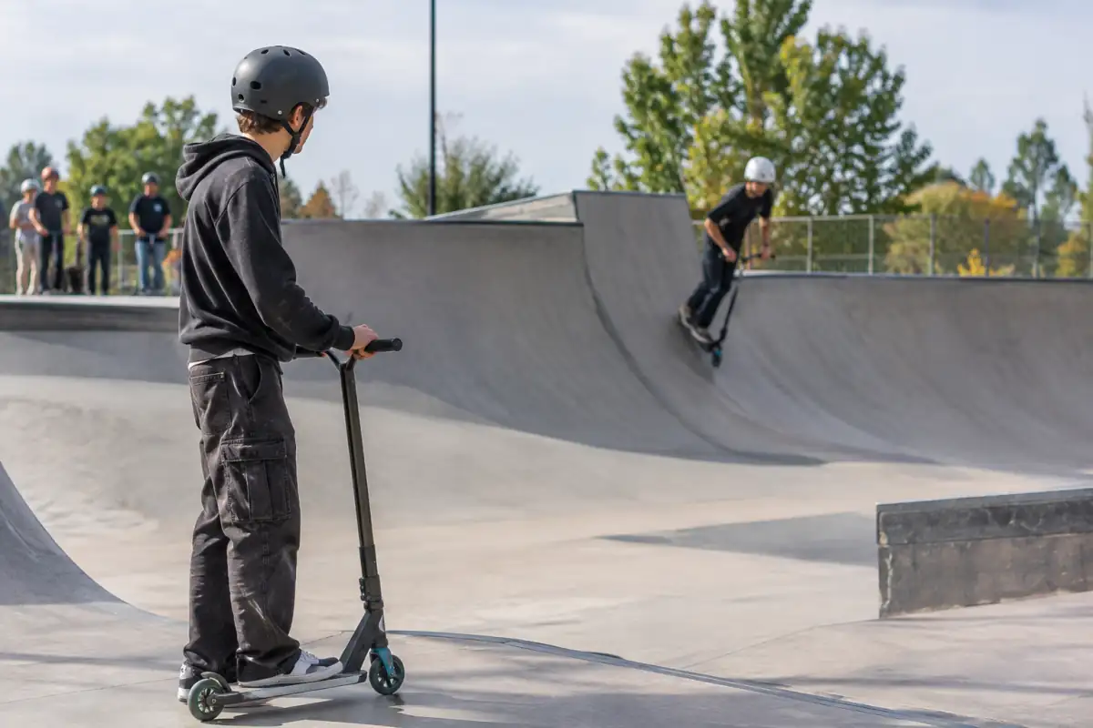 Dans un skatepark, un rider attend hors de la réception pendant qu’un autre effectue son passage en trottinette freestyle.