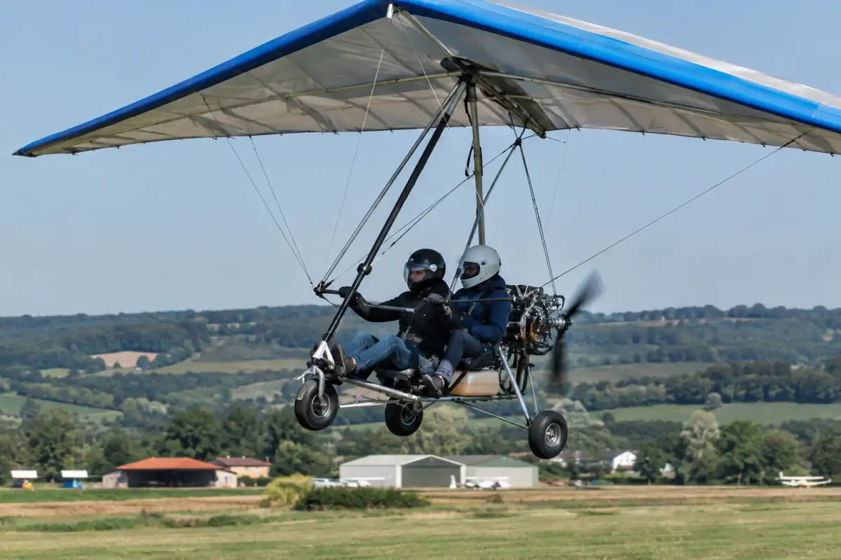 Un ULM pendulaire biplace vole au-dessus d’un aérodrome, son aile delta souple portée par un chariot motorisé.