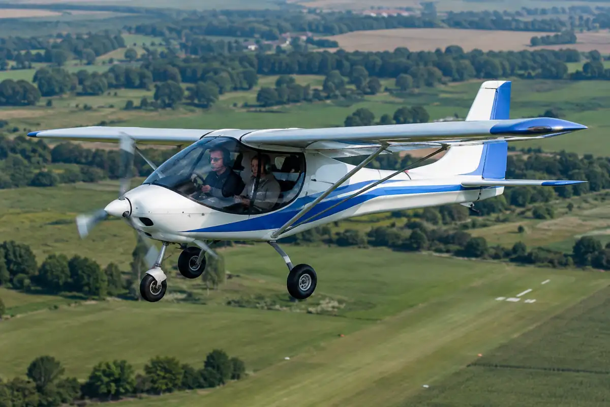 Un ULM multiaxes biplace en vol au-dessus d’un terrain d’aviation français, avec deux occupants équipés de casques.