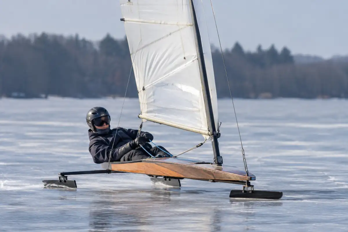 Un pilote en casque et vêtements chauds dirige un yacht DN à voile sur la glace d’un lac gelé.