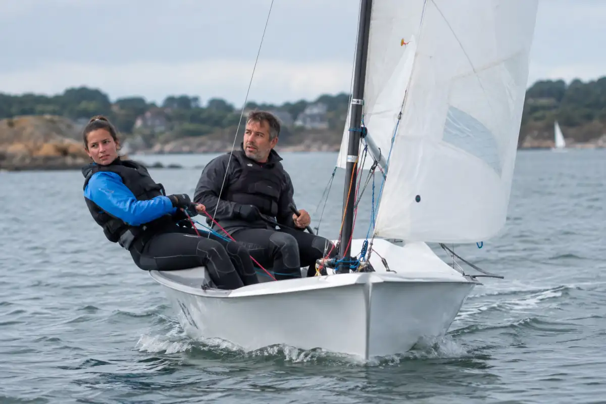 Deux débutants en voile règlent les voiles d’un dériveur légèrement incliné sur un plan d’eau côtier.