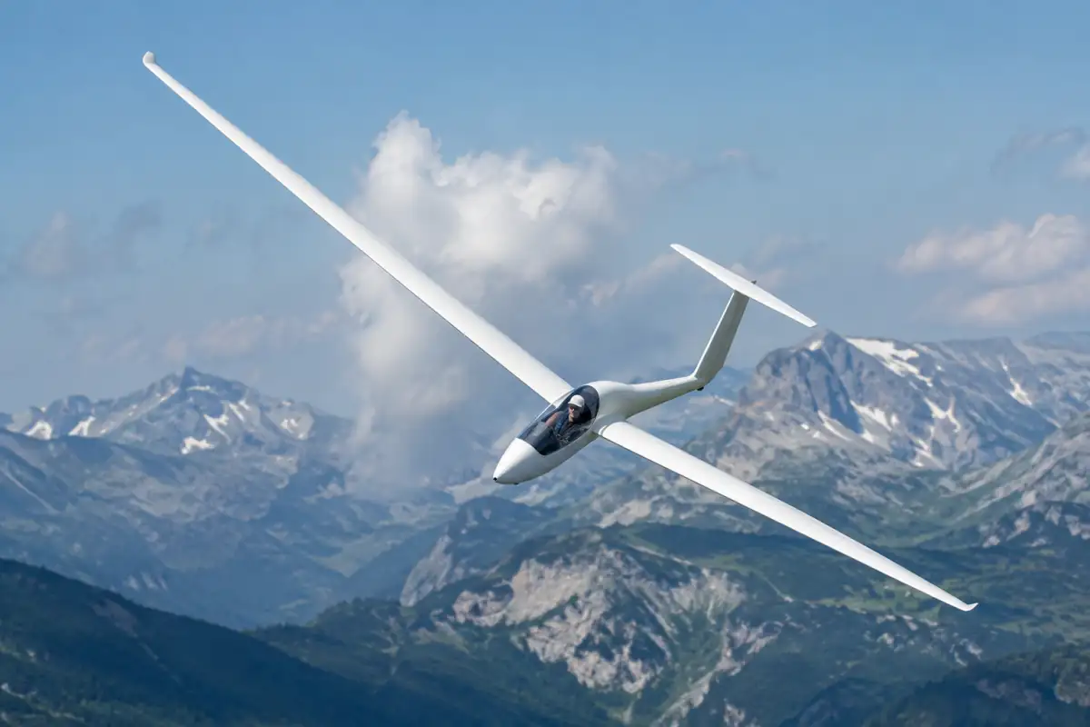 Un planeur monoplace en virage dans une ascendance, au-dessus d’un relief alpin parsemé de nuages.