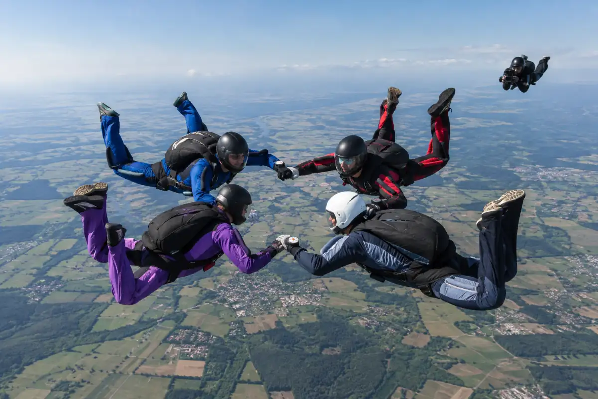 Quatre parachutistes en combinaison réalisent une formation de vol relatif en chute libre, filmés par un vidéoman.