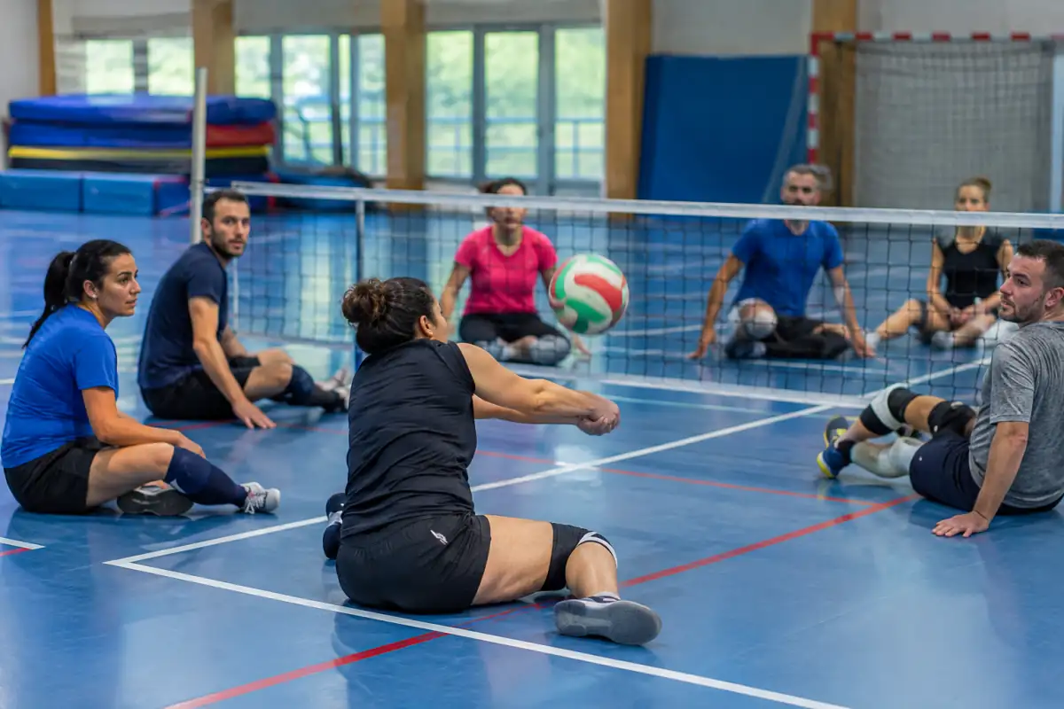 Échange de volley assis en salle : une joueuse réceptionne le ballon au sol tandis que ses coéquipiers se replacent.