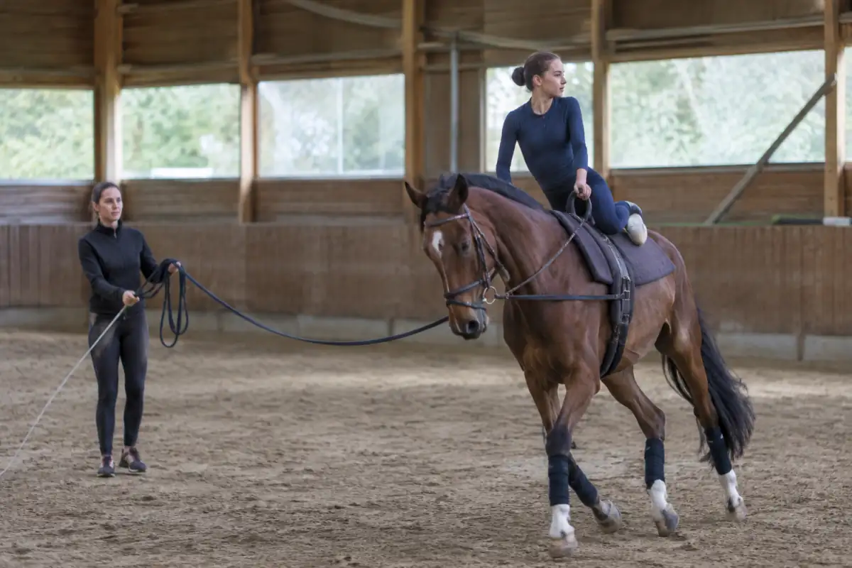 Une voltigeuse en équilibre à genoux sur un cheval équipé d’un surfaix, guidé au galop par un longeur dans un manège.