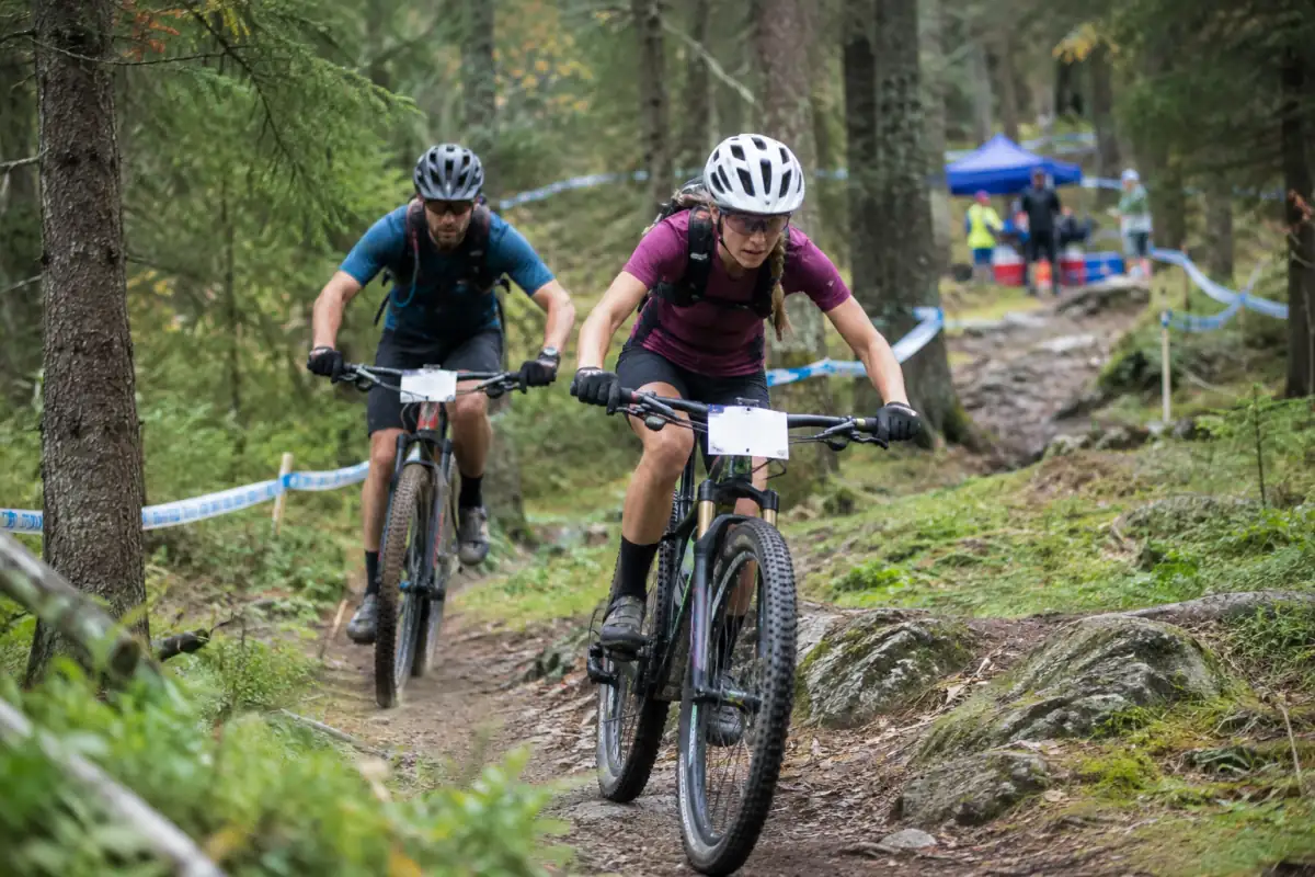 Deux vététistes en course sur un circuit balisé de cross-country, le second préparant un dépassement prudent sur une portion élargie.