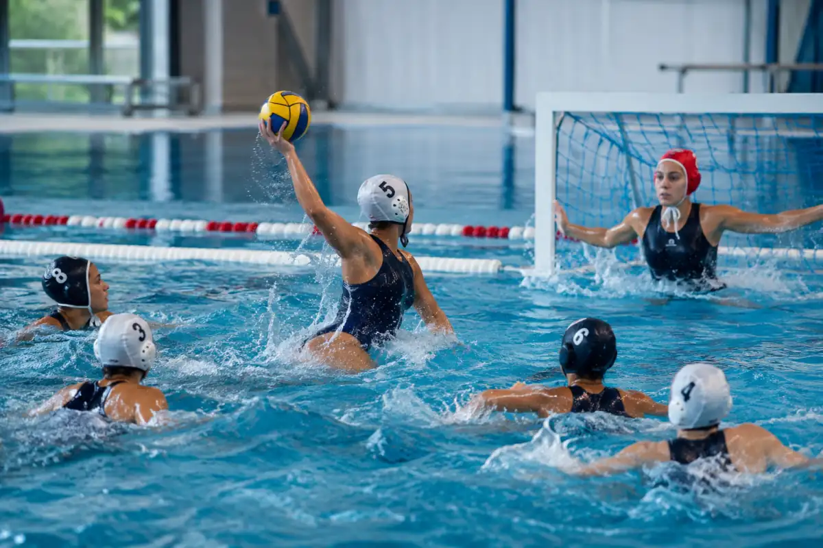 Joueur de water-polo s’élève hors de l’eau pour passer le ballon pendant un match en piscine, bonnet à protège-oreilles visible.