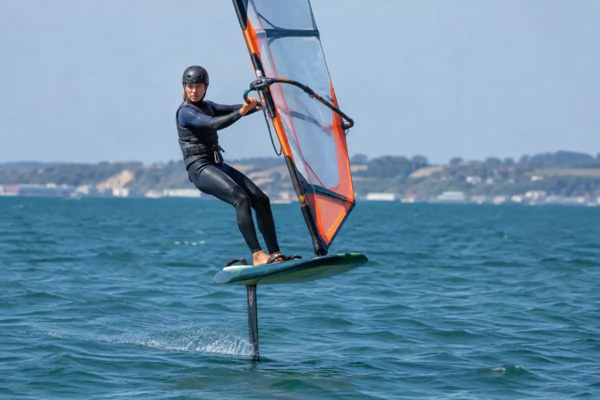 Un pratiquant de windfoil navigue au-dessus de l’eau, voile tenue au wishbone et foil visible sous la planche.