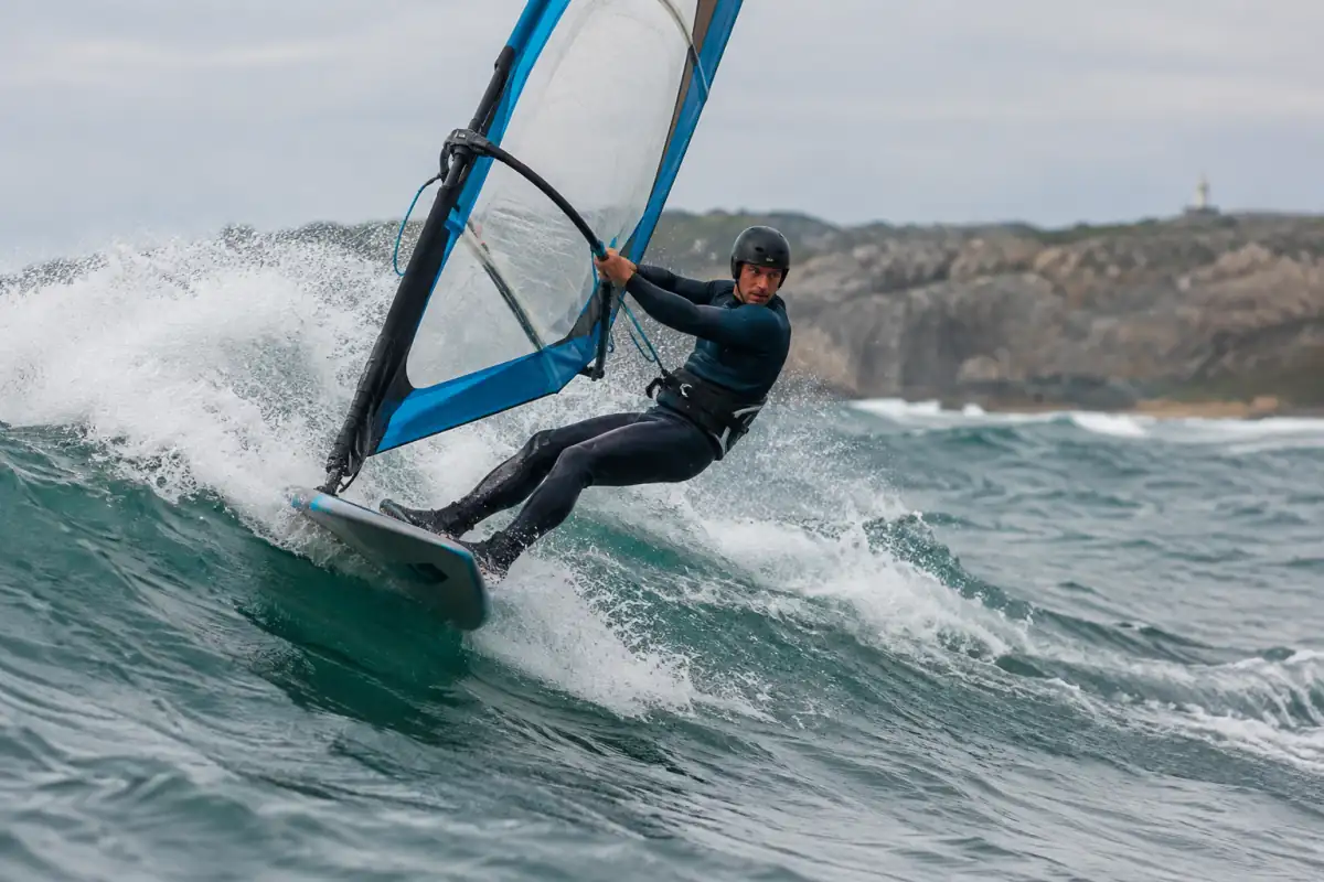 Véliplanchiste en combinaison et casque réalisant un virage sur la face d’une vague avec une voile de windsurf de vagues.
