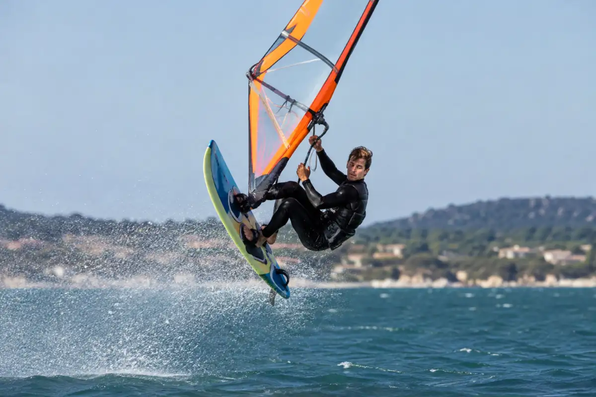 Un véliplanchiste réalise une rotation aérienne en windsurf freestyle au-dessus d’une eau plate, voile tenue à deux mains.