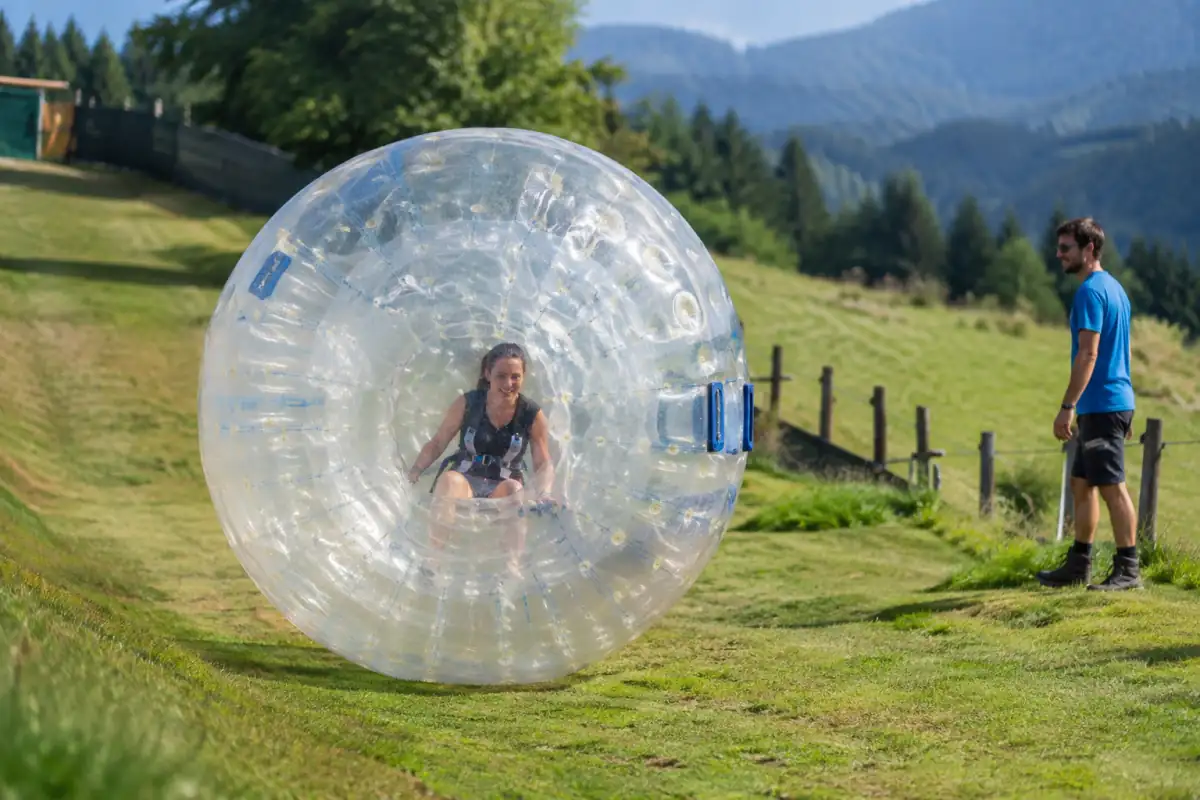 Une personne roule dans une sphère transparente sur une pente herbeuse aménagée, sous la surveillance d’un opérateur de zorbing.
